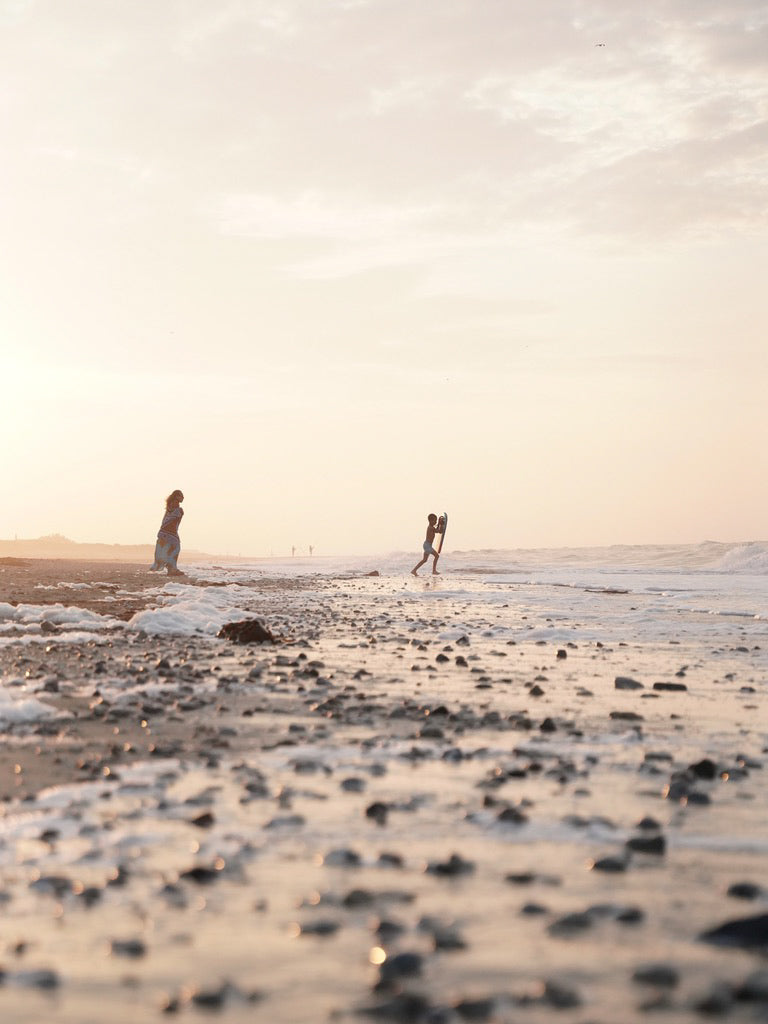 Valente Candles Glücksmoment: Mutter beobachtet Kind beim Wellenreiten am Meer im Sonnenuntergang.